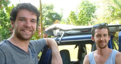 2 men in front of blue pick up with surfboard on top