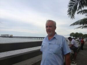 Older man in blue shirt standing by beach railing